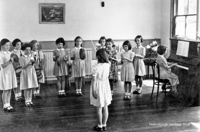 1950s St Bride's School class
A junior class at St Bride's School. From left: Val McIntyre, Sarah Lee, Fiona Carslaw or Pat Wright?, Vari Sandeman, Sandra McDougall, Barbara Brown, Alison Hamilton, Esther Henderson in floral dress, Phyllis Marshall, Liz Birnie, with Ann Davidson at the piano and Alison Gow conducting. Image circa 1957.
