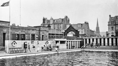 Helensburgh Swimming Pool
A 1959 image showing the entrance and the north west corner of the outdoor pool, built in 1928 as a gift from Provost Andrew Buchanan, beside Helensburgh Pier. It was replaced in 1976 by an indoor pool on an adjacent site, and a year later the outdoor pool was closed. It was demolished in 1996 and replaced by a childrens play area. Behind it is an archway which led to the pier, but was demolished some years ago.
