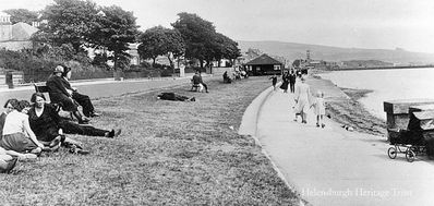 West Esplanade
Relaxing on the grass of the West Esplanade in Helensburgh, with big trees in the gardens of the seafront villas and one of the now demolished shelters in the distance. Image circa 1935.
